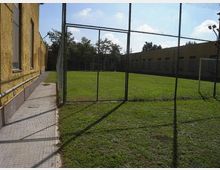 Campo da calcio in erba recintato con rete metallica, con porte bianche alle estremità. A sinistra corre un marciapiede lungo un edificio giallo e sullo sfondo si vedono altri edifici bassi e alberi sotto un cielo sereno.