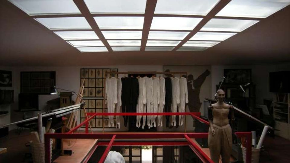 Interno di un atelier o laboratorio di sartoria con grande lucernario a griglia sul soffitto e passerella con ringhiera rossa attorno a un vuoto centrale. Sul fondo sono appesi capi chiari e scuri su una barra, con arredi e attrezzature da lavoro ai lati.