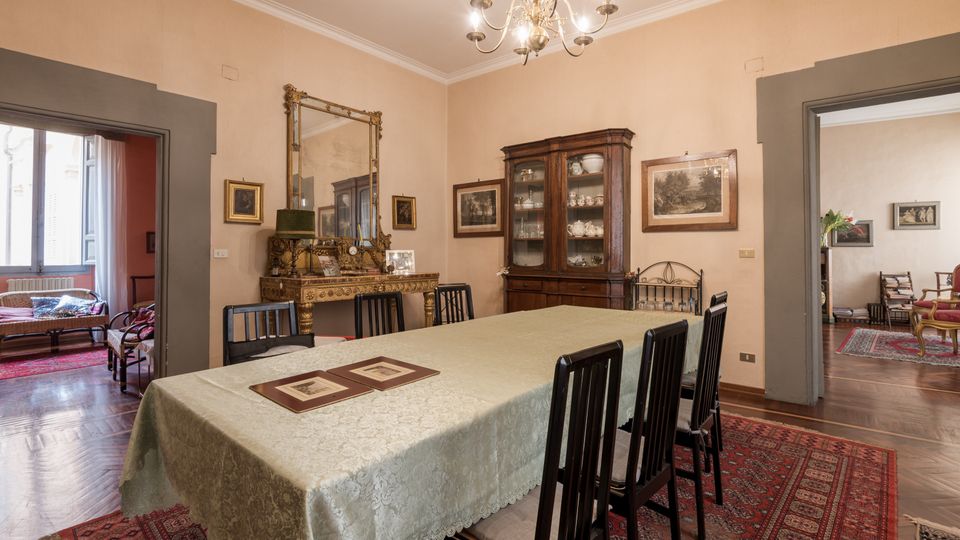 Sala da pranzo in stile classico con grande tavolo rettangolare coperto da tovaglia chiara, sedie scure e tappeti orientali su pavimento in legno. Alle pareti ci sono quadri e uno specchio sopra una consolle decorata, con lampadario a bracci sul soffitto e vetrina in legno; due ampie aperture collegano la stanza ad ambienti adiacenti.