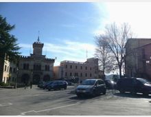 Piazza cittadina con ampio parcheggio e auto, affacciata su un edificio storico in stile municipale con portici ad arcate, merlature e torre centrale. Sul lato destro si vedono alberi spogli e altri edifici a più piani sotto un cielo sereno.
