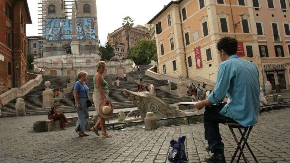 Piazza pavimentata in pietra con ampia scalinata monumentale che sale verso una chiesa con due campanili in restauro e teli protettivi. Ai lati si vedono palazzi storici con finestre e persiane, una fontana con vasca e balaustre in pietra.