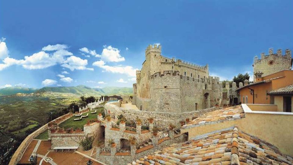 Veduta panoramica di un castello medievale in pietra con mura merlate e torri, affacciato su una terrazza con parapetti e vasi. In primo piano si vedono tetti in coppi e, sullo sfondo, una valle collinare verde sotto un cielo azzurro con nuvole.