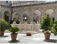 Cortile interno in pietra con loggiato ad archi e balaustra superiore, con al centro una fontana rettangolare in muratura. Pavimentazione in lastre di pietra e grandi vasi in terracotta con arbusti e fiori disposti intorno.