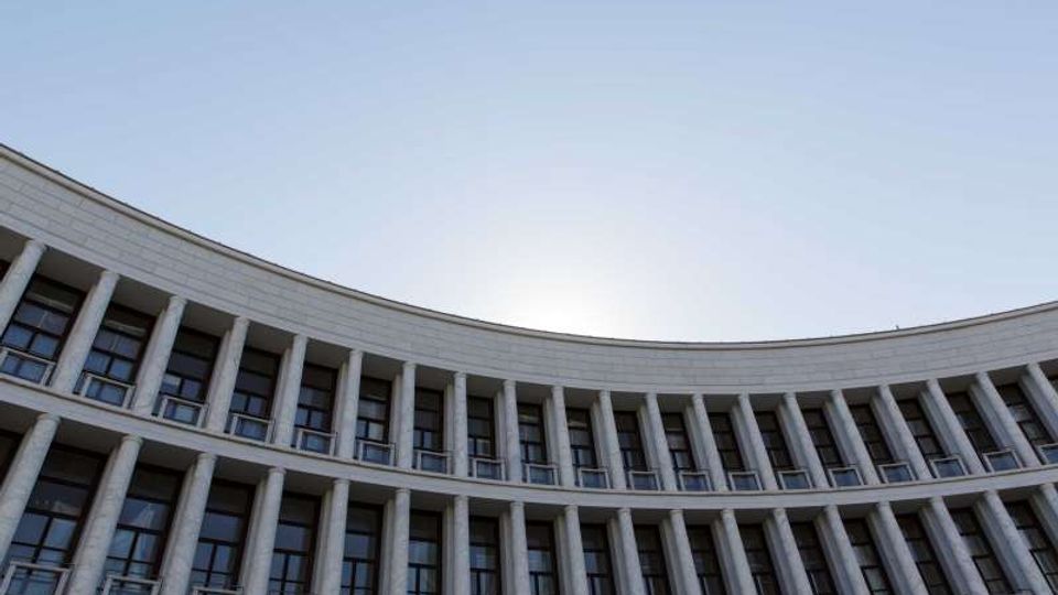 Facciata curva di un grande edificio moderno con file regolari di colonne verticali e finestre su più piani, ripresa dal basso verso l’alto. Sullo sfondo si vede il cielo limpido.