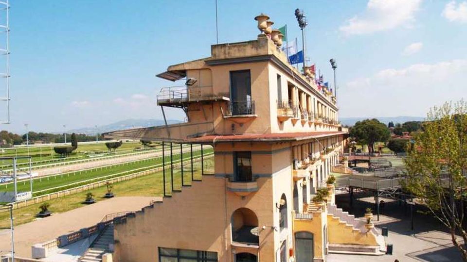Edificio principale di un ippodromo, a più piani, con facciata in intonaco chiaro, balconi e terrazze, affacciato su una pista di corsa con prato e ringhiere. Intorno si vedono gradinate, scale esterne, strutture tecniche e alberi, con cielo sereno sullo sfondo.