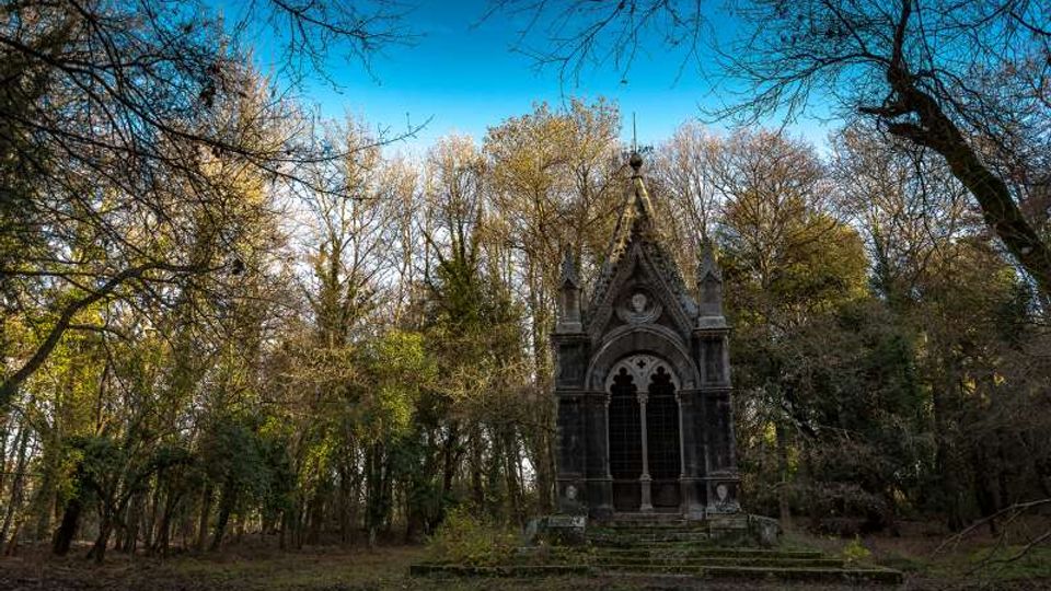 Piccolo mausoleo in pietra in stile gotico, con facciata a timpano e alte finestre ad arco, situato in una radura. Intorno si estende un bosco fitto di alberi alti e spogli, con cielo azzurro visibile tra i rami.