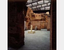 Interno di un’area archeologica coperta, con pavimento in cemento e pareti in mattoni antichi parzialmente crollate. Al centro si trova una piccola statua su basamento, sotto una grande copertura con travi metalliche e pannelli traslucidi.
