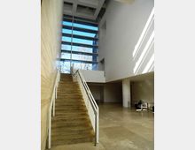 Atrio interno moderno con ampia scala in pietra e corrimano metallici, pavimento in grandi lastre e soffitto alto. Una grande vetrata a tutta altezza illumina lo spazio, con vista su alberi all’esterno.