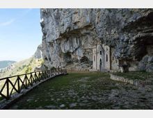 Piccola chiesa in pietra con facciata gotica incastonata sotto una grande parete rocciosa a strapiombo, accanto a una terrazza erbosa e pavimentata. Un camminamento con staccionata in legno segue il bordo della scogliera con vista sulle montagne circostanti.