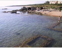 Costa rocciosa con piccole calette e acqua bassa e limpida, con scogli affioranti e piattaforme di roccia coperte di alghe. Sullo sfondo si vedono grotte nella falesia, vegetazione costiera e un edificio vicino alla scogliera.