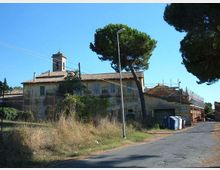 Edificio rurale a due piani con tetto in coppi e una piccola torre campanaria, circondato da recinzioni e vegetazione incolta. La scena è lungo una strada asfaltata con lampione, pini alti e alcuni cassonetti, sotto un cielo sereno.