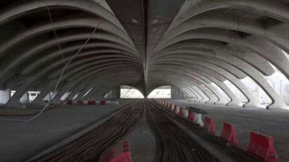 Interno di un grande viadotto o sottopasso in cemento con archi ripetuti e soffitto a volte, visto in prospettiva verso un’apertura luminosa. Sul pavimento si vedono fasci di cavi o barre stesi longitudinalmente e barriere stradali rosse e bianche ai lati.