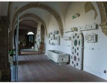 Corridoio interno con archi in pietra e soffitto a volta, pavimento in mattoni e pareti bianche su cui sono esposti frammenti architettonici e pannelli decorativi. Lungo la parete si vedono una vasca in pietra e vari reperti, con una porta ad arco in fondo al passaggio.