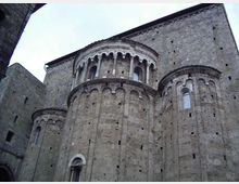 Vista esterna di una chiesa romanica in pietra con absidi semicircolari e decorazioni ad archetti ciechi. La facciata laterale mostra un alto tamburo con loggiato ad arcate e piccole finestre, su muratura massiccia in blocchi di pietra.