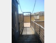 Passerella in legno con corrimano metallici e catene laterali, che conduce a un’uscita rettangolare verso un cortile pavimentato in mattoni. Sullo sfondo ci sono muretti in cemento, una breve scalinata e edifici lontani sotto un cielo sereno.