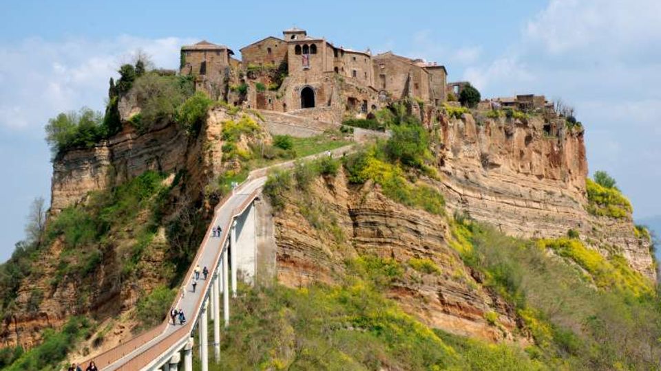 Borgo storico in pietra arroccato sulla sommità di una rupe di tufo, con case e una chiesa al centro. Un lungo ponte pedonale sopraelevato conduce all’ingresso, circondato da pendii verdi e stratificazioni rocciose sotto un cielo parzialmente nuvoloso.