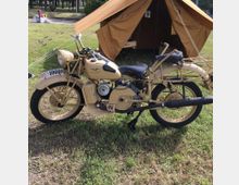 Motocicletta d’epoca color beige parcheggiata su un prato, con cavalletto e ruote a raggi in evidenza. Sullo sfondo si vede una tenda da campo in tela marrone montata in un’area verde, con alberi e altre strutture lontane.