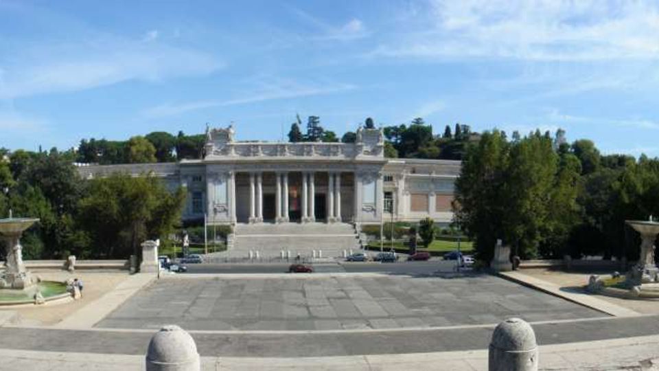 Vista panoramica di una grande piazza pavimentata con due fontane simmetriche ai lati, che conduce a un edificio monumentale in stile neoclassico con ampia scalinata e colonnato centrale. Sullo sfondo si vedono alberi e una collina verde sotto un cielo azzurro.