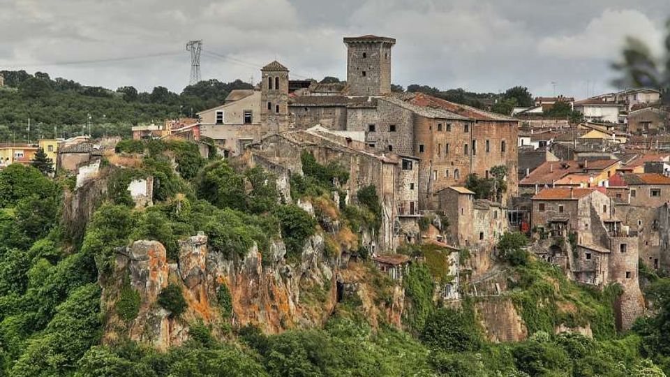 Borgo storico in pietra arroccato su una rupe, con case addossate e un grande edificio fortificato con torre quadrata e campanile. In primo piano si vedono le pareti rocciose e la vegetazione, con colline e cielo nuvoloso sullo sfondo.