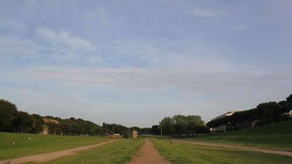 Ampio prato in un parco con un sentiero sterrato centrale che conduce verso edifici lontani all’orizzonte. Ai lati si vedono alberi e lievi pendii erbosi sotto un grande cielo azzurro con nuvole sottili.