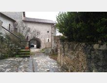 Cortile in pietra con pavimentazione acciottolata che conduce a un arco d’ingresso in un edificio in muratura, con tetto a coppi. Ai lati si vedono un muretto in pietra con vegetazione fitta e una scala esterna con ringhiera metallica.