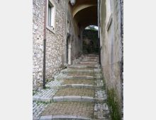 Vicolo stretto in pietra con pavimentazione a ciottoli e gradini in mattoni e lastre, tra muri di edifici antichi. In fondo si vede un passaggio ad arco coperto con una lanterna e una breve rampa che sale.