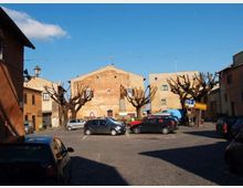 Piazza di un piccolo centro con parcheggio asfaltato e auto in sosta, circondata da edifici in muratura dai toni ocra. Al centro si vede una statua su piedistallo e una fila di alberi potati senza foglie sotto un cielo sereno.