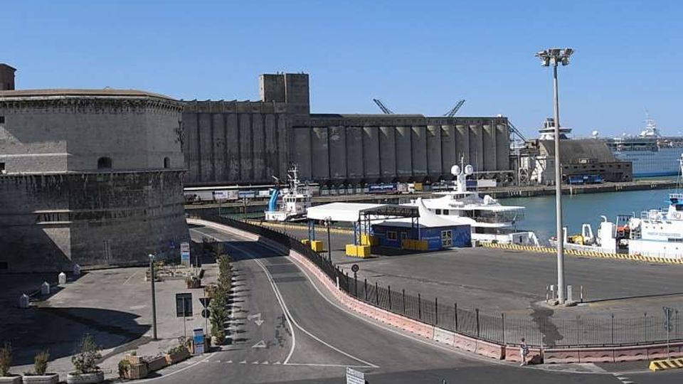 Veduta di un porto industriale con banchine e imbarcazioni ormeggiate, affiancato da grandi edifici in cemento tipo silos e magazzini. In primo piano si vedono una strada curvilinea con incrocio, recinzioni e lampioni lungo l’area portuale.