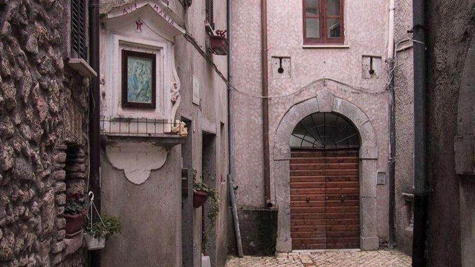 Vicolo stretto in un borgo storico con pavimentazione in pietra, muri in muratura grezza e intonaco, e cavi a vista. In fondo si vede un grande portone ad arco in pietra con porta in legno, una lanterna sporgente e vasi di piante lungo le pareti.