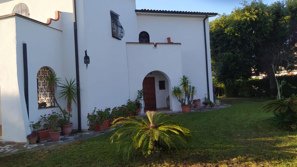 Esterno di una casa in stile mediterraneo con pareti bianche e tetto in tegole, ingresso ad arco e finestra con grata in ferro battuto. Davanti c’è un prato con piante in vaso e una piccola palma, con alberi e vegetazione sullo sfondo.