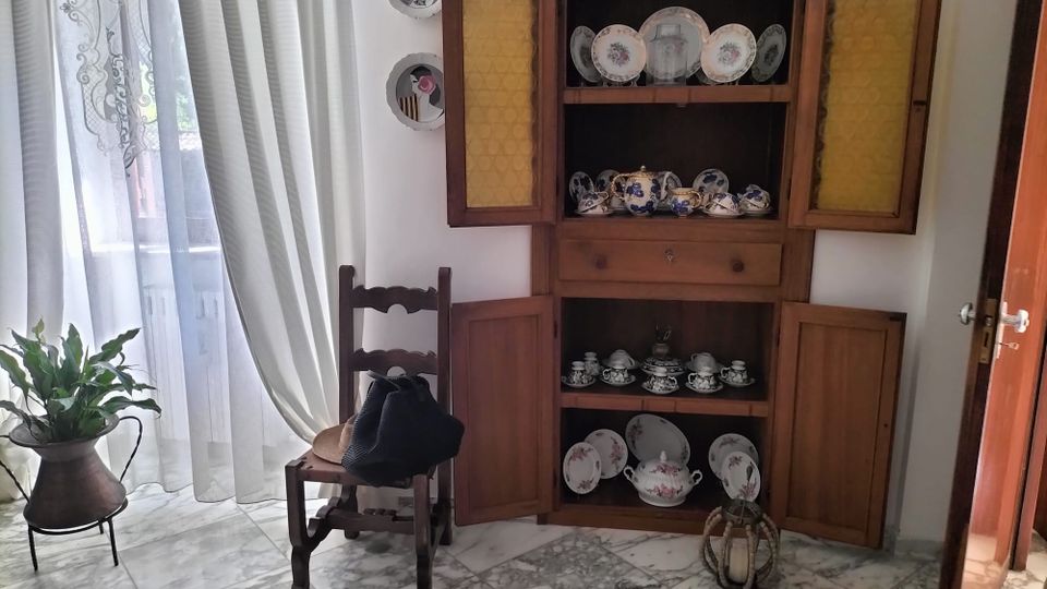 Angolo di soggiorno con pavimento in marmo e grande finestra con tende chiare che lascia entrare luce naturale. Accanto a una sedia in legno e a un vaso con pianta, c’è una credenza in legno con ante aperte che espone piatti e servizi da tè in ceramica.