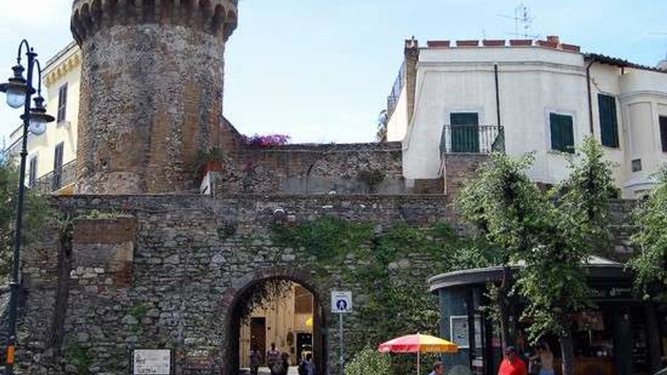 Ingresso ad arco in una cinta muraria in pietra, con una torre cilindrica merlata in stile medievale e vegetazione rampicante sulle mura. Sullo sfondo si vedono edifici chiari con persiane verdi e, in primo piano, una piccola area urbana con lampioni, alberi e un chiosco.