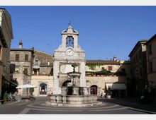 Piazza storica con una fontana in pietra al centro e, sullo sfondo, una facciata monumentale con campanile e orologio. Intorno si vedono edifici antichi a più piani con finestre e persiane, sotto un cielo sereno.