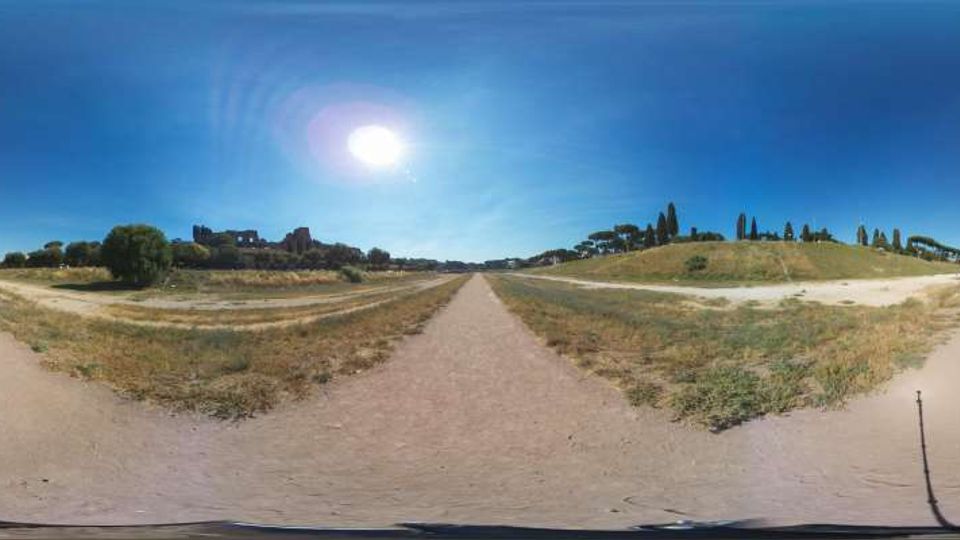 Vista panoramica di un ampio viale sterrato in un’area archeologica, con prati secchi ai lati e colline basse. Sullo sfondo si vedono rovine in muratura, filari di cipressi e un cielo azzurro con il sole alto.