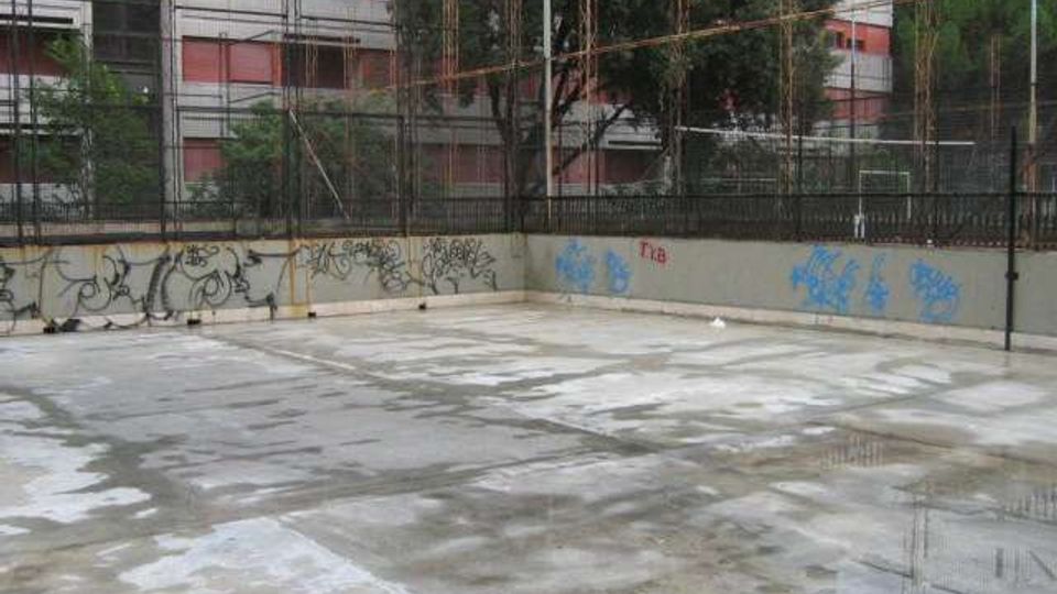 Cortile o area pavimentata in cemento con pozzanghere, circondata da muri bassi con graffiti e una recinzione metallica. Sullo sfondo si vedono alberi e un edificio residenziale a più piani con balconi e tende rosse.