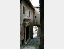 Vicolo stretto in pietra tra edifici in muratura intonacata, con portale ad arco in pietra lavorata e piccole finestre rettangolari. In fondo, un arco passante conduce a una stradina acciottolata e a uno spazio esterno illuminato.