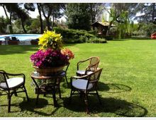 Giardino con ampio prato verde e area salotto con tavolino e sedie in vimini, con un cesto di fiori colorati al centro. Sullo sfondo si vedono una piscina, alberi alti e una piccola struttura con tetto in tegole tra la vegetazione.