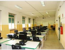 Aula informatica con file di banchi dotati di computer e monitor, pareti bianche e gialle e illuminazione a neon sul soffitto. Lungo un lato ci sono finestre con tapparelle verdi e, in fondo, una lavagna con proiettore a soffitto.