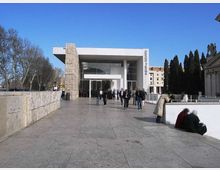 Ingresso di un edificio moderno con ampia facciata vetrata e tetto piano aggettante, affiancato da un pilastro rivestito in pietra e con la scritta verticale “fabrizio de andrè”. Davanti si apre una grande piazza pavimentata in pietra, con muretto laterale, alberi spogli e altri edifici sullo sfondo.