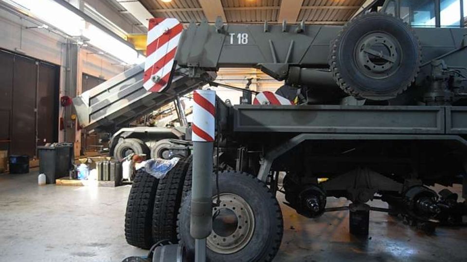 Interno di un’officina o capannone industriale con pavimento in cemento e tetto in metallo illuminato da luci a soffitto. In primo piano si vede un grande veicolo gru in manutenzione, con braccio e stabilizzatori con bande rosse e bianche, ruote e componenti meccanici sparsi a terra.