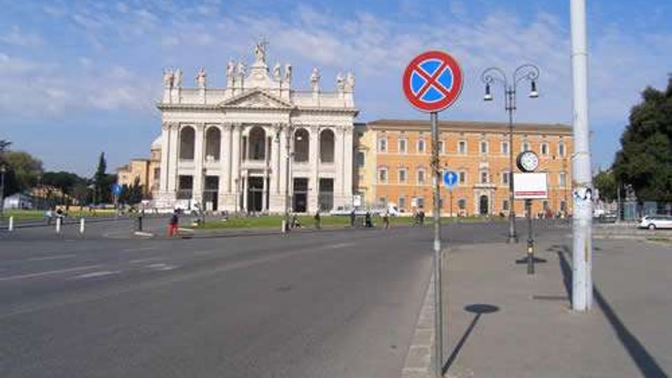 Ampia piazza urbana con strada asfaltata e marciapiede in primo piano, segnaletica di divieto di sosta e lampioni. Sullo sfondo si vede una grande basilica in stile neoclassico con facciata bianca a colonne e statue, affiancata da edifici storici color ocra.