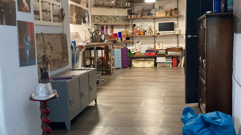 Interno di uno studio creativo con pavimento in legno grigio e un piccolo gradino d’ingresso; pareti bianche con foto e stampe, scaffali a vista pieni di materiali e un’area di lavoro illuminata da lampade a sospensione. Sul lato destro si vede una porta scura e un armadio, mentre in primo piano ci sono un pouf blu e un piccolo supporto rosso.