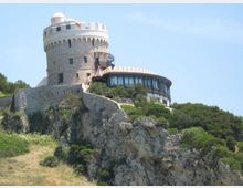 Torre fortificata in pietra con merlature, situata su un promontorio roccioso e collegata a un edificio semicircolare con grandi vetrate. Intorno si vede vegetazione mediterranea su pendii e scogli, sotto un cielo sereno.