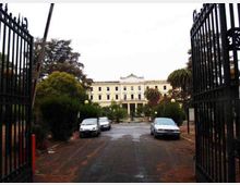 Ingresso con cancelli in ferro battuto aperti su un viale asfaltato che conduce a un grande edificio neoclassico a più piani, con facciata chiara e portico centrale. Ai lati del viale ci sono alberi e siepi, lampioni e alcune auto parcheggiate.