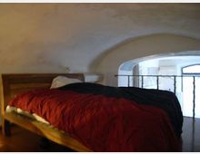 Camera da letto in un soppalco con soffitto a volta intonacato e letto in legno con coperta rossa e nera. Un’apertura ad arco con ringhiera in ferro affaccia su una zona sottostante illuminata da una finestra.