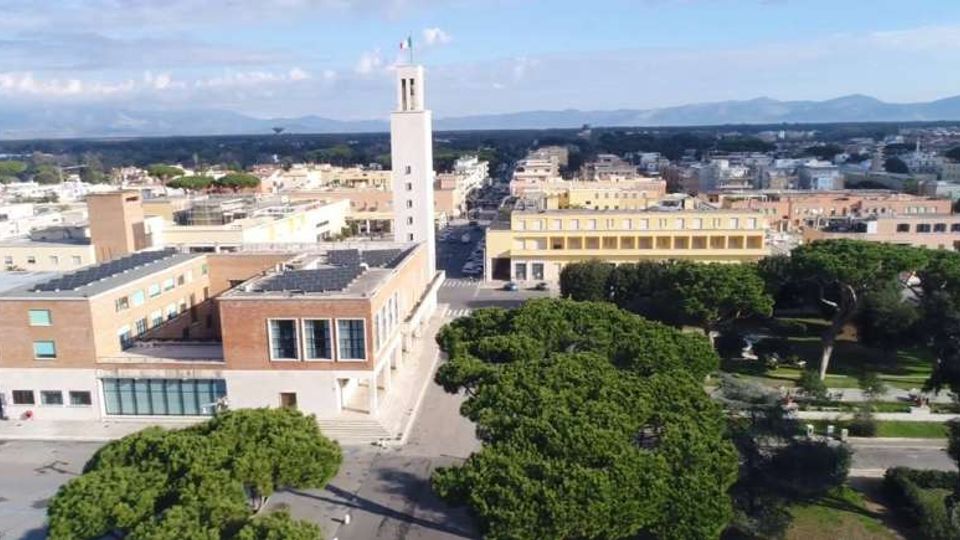 Veduta aerea di un complesso civico moderno con torre bianca centrale e edifici bassi in mattoni e intonaco, affacciati su una piazza. In primo piano un parco con alberi e vialetti, sullo sfondo la città e una catena di montagne.