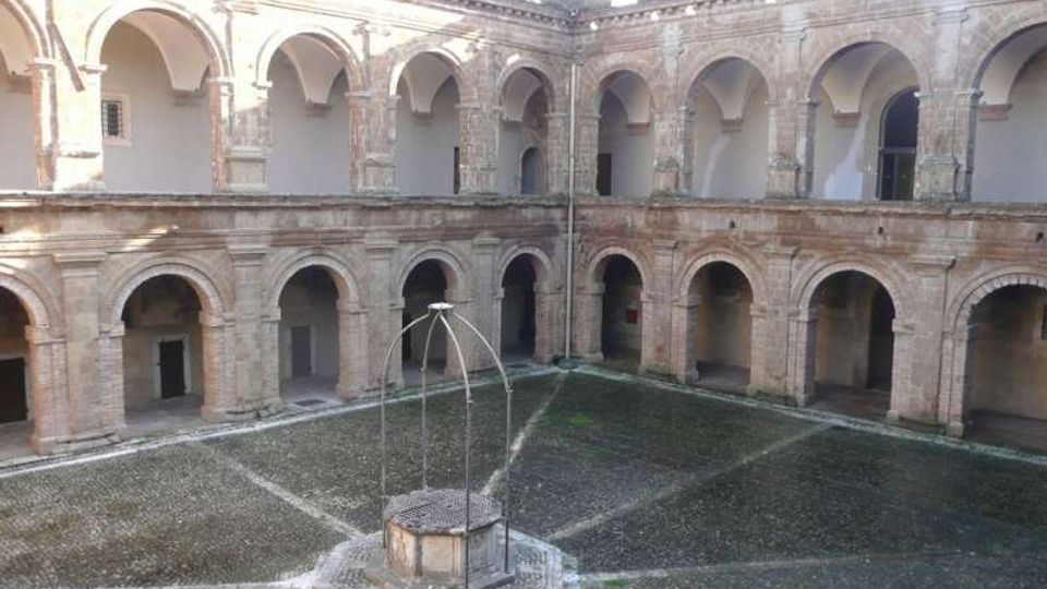 Cortile interno di un edificio storico in pietra, circondato da un porticato su due livelli con archi e colonne. Al centro si trova un pozzo con struttura metallica, su pavimentazione in ciottoli disposta a raggiera.