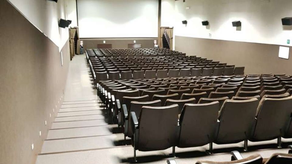 Sala auditorium con file di poltrone disposte in gradinata e corridoi laterali, rivolta verso uno schermo bianco frontale. Pareti bicolori con altoparlanti montati e soffitto scuro con luci a pannello incassate.