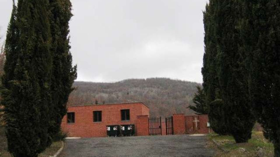 Ingresso asfaltato fiancheggiato da alti cipressi che conduce a un edificio basso in mattoni rossi con finestre rettangolari e un cancello metallico. Sullo sfondo si vedono colline boscate sotto un cielo coperto.
