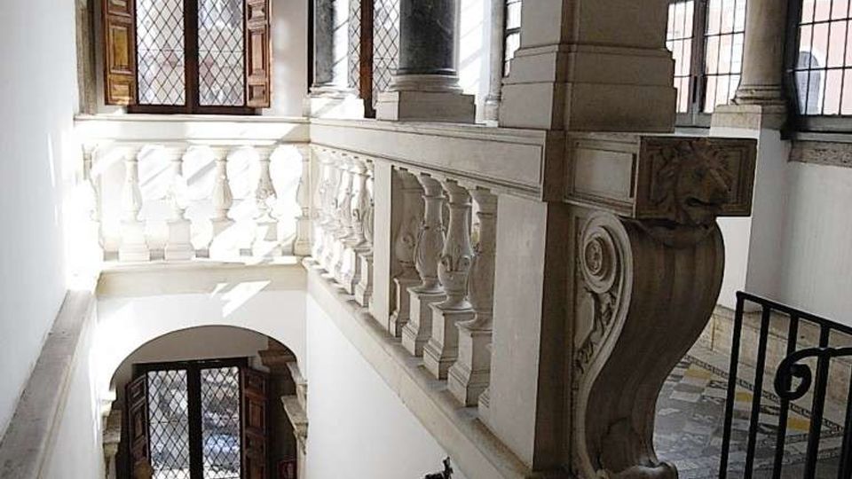 Interno di un edificio storico con scala in pietra che scende verso un pianerottolo, affiancata da una balaustra in marmo scolpito. Grandi finestre con griglie e colonne in marmo illuminano l’ambiente, con soffitto decorato e pavimento a mosaico visibili al piano superiore.